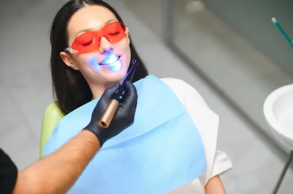 A female patient undergoing a dental bonding procedure while wearing protective red glasses, with a curing light being applied to her teeth.