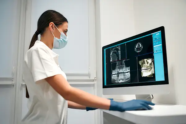 Dental professional in a mask and gloves examining high-resolution dental images on a computer for full-mouth restoration planning.